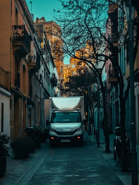 A white Iveco van parked on a narrow urban street between multi-storey residential buildings with balconies and ornate window details. The van is positioned centrally and is partially loaded with larg